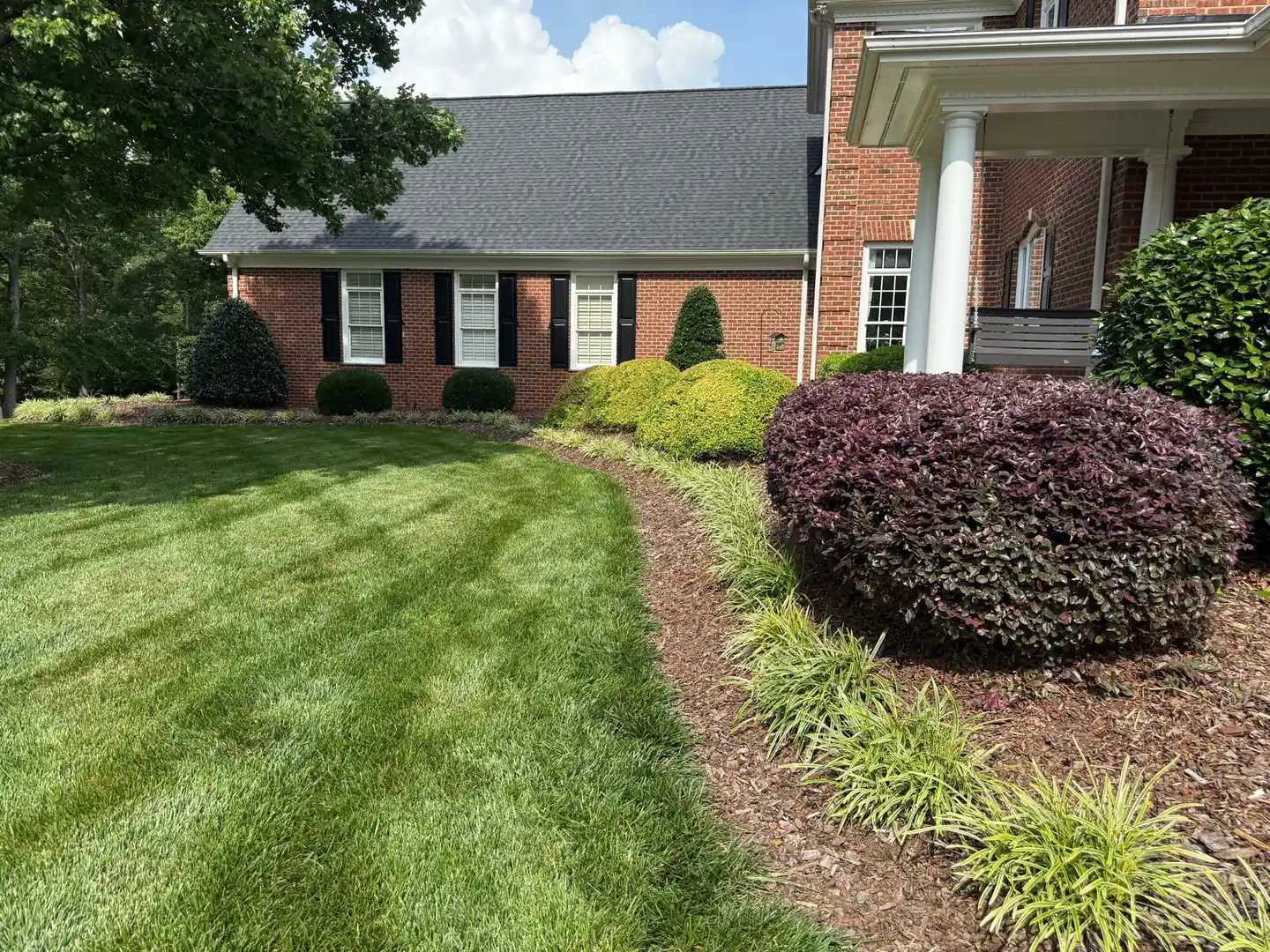 A brick house with dark roof shingles, a green lawn, and various shrubs in a landscaped yard on a sunny day.